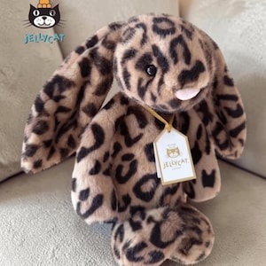 Op de afbeelding: Een pluche konijn met een luipaardprint. Het konijn heeft lange, slappe oren, een roze neus en een label met de tekst "Jellycat". Het speelgoed is bruin en zwart en ligt op een grijze ondergrond.