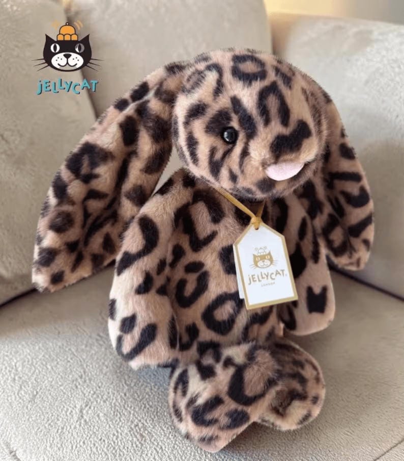 Op de afbeelding: Een pluche konijn met een luipaardprint. Het konijn heeft lange, slappe oren, een roze neus en een label met de tekst "Jellycat". Het speelgoed is bruin en zwart en ligt op een grijze ondergrond.