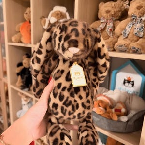 Op de afbeelding: Een pluche konijn met luipaardprint, lange slappe oren en een roze neus. Het knuffel heeft een label met de tekst "Jellycat". Andere knuffels zijn zichtbaar op planken op de achtergrond.