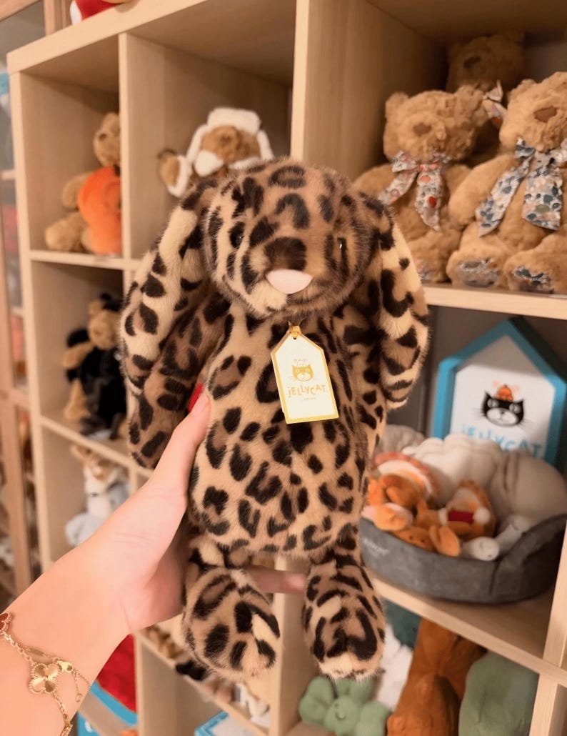 Op de afbeelding: Een pluche konijn met luipaardprint, lange slappe oren en een roze neus. Het knuffel heeft een label met de tekst "Jellycat". Andere knuffels zijn zichtbaar op planken op de achtergrond.
