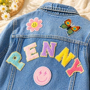May include: A blue denim jacket featuring colorful patches on the back. The patches include a pink flower with a smiley face, a multi-colored butterfly, and a pink smiley face with lightning bolt eyes. The name PENNY is spelled out in large letters.