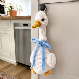 May include: A white crochet goose with a yellow beak and feet, and black eyes. The goose has a light blue bow around its neck. The handmade goose is hanging on a door.