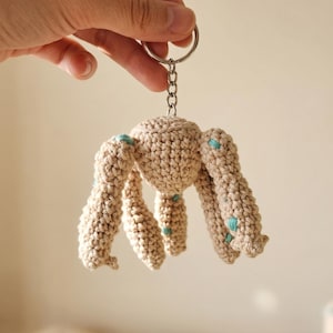 Puede incluir: Un llavero de ganchillo con una criatura beige con forma de pulpo y detalles azules. El amuleto hecho a mano tiene un cuerpo redondo y múltiples patas texturizadas. El llavero está sujeto a un anillo de metal plateado.