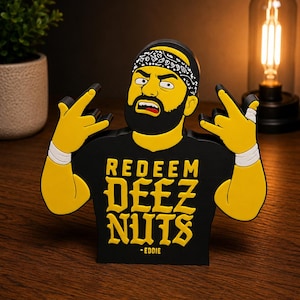 May include: A black and yellow figurine of a man with a beard and bandana, making a hand gesture. The figurine has the text "REDEEM DEEZ NUTS - EDDIE" on the front. The figurine is on a wooden surface.