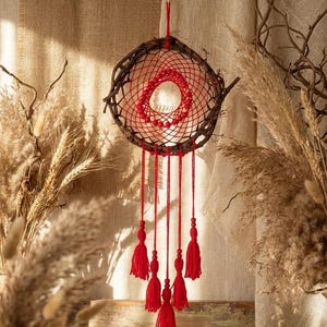 Puede incluir: Un atrapasueños artesanal con un marco circular tejido con ramitas. El interior presenta un diseño de red roja con una abertura central y borlas rojas colgando. El fondo incluye plantas secas y tela texturizada.