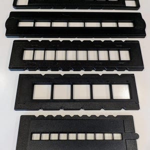 Puede incluir: Conjunto de cinco monturas de diapositivas de película negras, cada una con aberturas rectangulares para sujetar tiras de película. Las monturas varían en diseño, con diferentes números de aberturas y detalles de borde. Las diapositivas están dispuestas verticalmente sobre un fondo blanco.