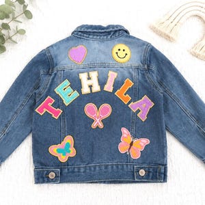 May include: A blue denim jacket featuring colorful patches on the back, including the name TEHILA in large block letters, a yellow smiley face, a purple heart, two pink tennis rackets, and two butterflies in shades of pink, yellow, and blue.