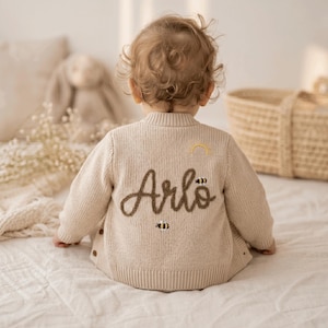 Könnte beinhalten: Beige Baby-Strickjacke mit dem Namen "Arlo" in brauner Schrift auf dem Rücken gestickt, zusammen mit einem kleinen Regenbogen und einem Bienen-Design. Die Strickjacke hat Knopfverschlüsse und ist für ein Baby konzipiert.