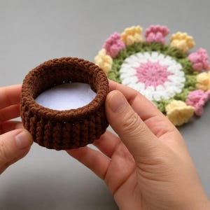 Puede incluir: Una pequeña cesta redonda de ganchillo marrón con interior blanco. La cesta se sostiene con las manos, con un posavasos de ganchillo colorido en el fondo. El posavasos presenta diseños en forma de flor rosa, amarilla y blanca.
