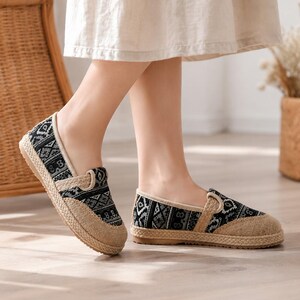 May include: A pair of slip-on shoes featuring a black and white geometric patterned fabric upper, a tan jute-textured toe cap, and a braided jute midsole. The shoes have a beige fabric trim along the collar and a flat, flexible rubber outsole.