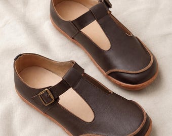 May include: A pair of dark brown T-strap shoes featuring a rounded toe, a side buckle closure with a metallic bronze-toned buckle, and a light tan interior lining. The shoes have a visible stitched edge along the sole and a smooth leather-like finish.