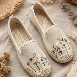 Può includere: Un paio di scarpe slip-on beige con fiori di campo ricamati nei toni del giallo, viola e marrone. Le scarpe presentano una punta in stile iuta intrecciata, una tomaia in tessuto testurizzato e una linguetta rinforzata sul tallone.