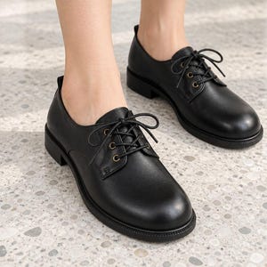May include: A pair of black leather oxford shoes featuring a rounded toe, a low block heel, and a lace-up closure with dark metal eyelets. The shoes have a smooth finish and a structured silhouette, providing a classic look with a durable rubber sole.