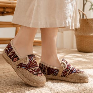 Puede incluir: Un par de zapatos sin cordones con una parte superior de tela roja oscura con un patrón geométrico multicolor, un ribete beige y un material texturizado estilo yute que cubre la puntera y el borde de la suela, creando un diseño casual.