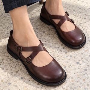 May include: A pair of dark brown leather shoes featuring a rounded toe and two adjustable crisscross straps across the top, each secured with a small metal buckle. The shoes have a flat black sole and a smooth, matte finish on the leather.
