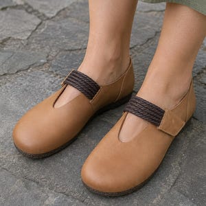 May include: A pair of tan leather flats featuring a rounded toe, a low-cut vamp, and a dark brown braided strap across the instep. The shoes have a flat sole with a dark brown braided trim detail along the edge, providing a simple and clean look.
