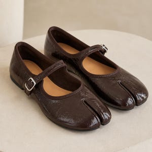 May include: A pair of dark brown, glossy patent leather Mary Jane flats featuring a distinctive split-toe design. Each shoe has a thin strap across the instep secured with a small silver-toned metal buckle and a dark brown interior lining.