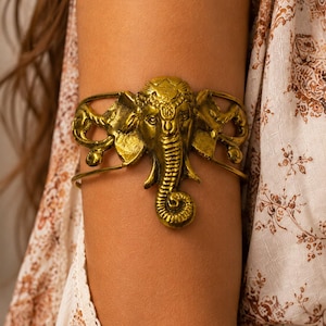 May include: A gold-toned metal arm cuff featuring a central elephant head motif with detailed trunk and ear textures. The band consists of two thin, curved metal wires that extend from the sides of the elephant head to form a circular shape.