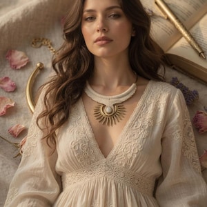 May include: A woman wearing a cream-colored dress with a deep V-neck and intricate embroidery. She is wearing a gold-toned necklace with a sunburst pendant. A gold pen and open book are in the background, along with rose petals.