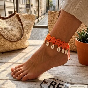 May include: A bright orange crocheted anklet featuring a scalloped edge design adorned with small white cowrie shells hanging from the bottom. The anklet is secured with a simple tie closure and is worn with a thin gold-toned metal toe ring.