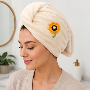 Pode incluir: Uma toalha de cabelo de microfibra na cor creme com uma superfície texturizada e felpuda. A toalha está enrolada num estilo torcido e decorada com uma pequena aplicação de girassol em croché com pétalas amarelas, centro castanho e caule verde.