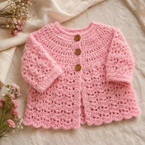 May include: A light pink crochet baby cardigan with three wooden buttons. The cardigan has a scalloped hem and three-quarter sleeves. The crochet pattern is open and airy, perfect for layering. The cardigan is displayed on a cream-colored surface.