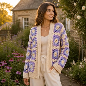 May include: A cream-colored, open-front crochet cardigan with a geometric pattern in lavender and yellow. The cardigan is worn over a cream-colored top and pants. The image is taken outdoors with a blurred background of a house and garden.