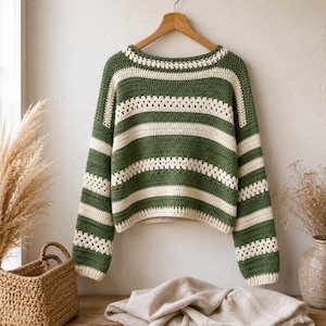 Puede incluir: Un suéter de ganchillo verde y crema con rayas horizontales cuelga de una percha de madera. El suéter tiene un cuello redondo y mangas largas. El fondo incluye una cesta, plantas secas y una pared de color neutro.