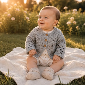 May include: A baby in a light grey crocheted cardigan with wooden buttons, a white onesie, and matching booties. The baby sits on a cream blanket on grass, with flowers in the background. The cardigan is a delicate, handmade item.