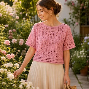 Könnte beinhalten: Ein rosa Häkeltop mit kurzen Ärmeln und einem runden Ausschnitt. Das Oberteil hat ein offenes, strukturiertes Design. Die Person trägt einen cremefarbenen Faltenrock. Der Hintergrund zeigt einen Garten mit rosa und weißen Blumen.