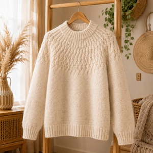May include: Cream-colored knit sweater with a round neckline and long sleeves. The upper chest area features a textured diamond pattern. The sweater is hanging on a wooden hanger against a neutral background, suggesting a cozy, handmade garment.