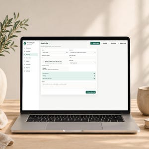 Pode incluir: Um laptop exibindo uma interface "Stock In" com detalhes do produto. A tela mostra campos para quantidade, data e outras informa&ccedil;&otilde;es de invent&aacute;rio. Uma caneca bege, um livro e um vaso decorativo com vegeta&ccedil;&atilde;o est&atilde;o na mesa de madeira.