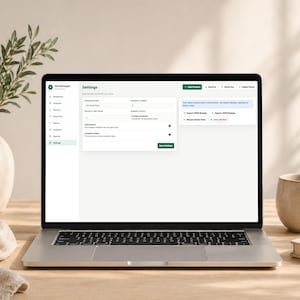 Pode incluir: Um laptop exibindo a p&aacute;gina "Configura&ccedil;&otilde;es" de um aplicativo de software. A tela mostra v&aacute;rias op&ccedil;&otilde;es de configura&ccedil;&otilde;es, incluindo contagens de produtos e recursos de exporta&ccedil;&atilde;o de dados. Uma caneca bege, um livro e um vaso decorativo com vegeta&ccedil;&atilde;o est&atilde;o na mesa de madeira.
