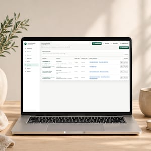 Pode incluir: Um laptop exibindo a p&aacute;gina "Fornecedores" da plataforma de e-commerce StockSimple. A tela mostra uma lista de fornecedores. Uma caneca, um livro e um vaso com vegeta&ccedil;&atilde;o est&atilde;o na mesa de madeira.