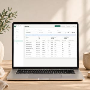 Pode incluir: Um laptop exibindo uma p&aacute;gina de "Relat&oacute;rios" com dados de invent&aacute;rio. A tela mostra detalhes do produto, quantidades e valores. Uma caneca bege e um livro est&atilde;o &agrave; direita. Um vaso com vegeta&ccedil;&atilde;o e um pano dobrado est&atilde;o &agrave; esquerda.