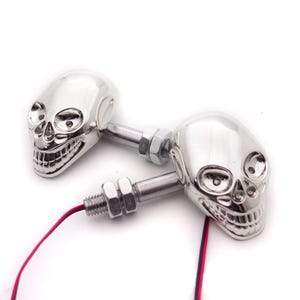 May include: Two chrome-finished skull-shaped motorcycle turn signal lights. Each skull features detailed eye sockets and teeth, attached to a threaded metal mounting bolt with two hexagonal nuts and a pair of red and black electrical wires.