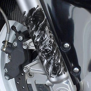 Può includere: Primo piano di una forcella anteriore di moto con un dettagliato design a teschio e tribale in bianco e nero. Il design è applicato alla forcella in metallo argentato, con altri componenti della moto visibili sullo sfondo.
