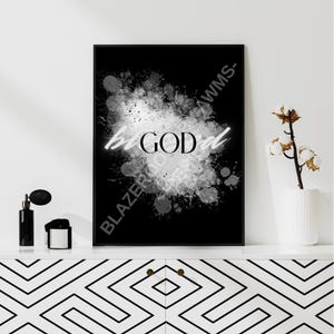 Könnte beinhalten: Schwarzer gerahmter Kunstdruck mit den Worten "be GOD" in weißer Neon-Schrift über einem weißen und grauen Spritzmuster. Das Kunstwerk wird auf einer weißen Oberfläche mit dekorativen Gegenständen und einem schwarz-weiß gemusterten Schrank präsentiert.
