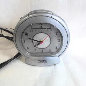 May include: A silver Timex AM/FM clock radio with an analog clock face featuring black hour and minute hands, a red second hand, and black numerals from 1 to 12. The device includes a radio frequency tuner scale above the center and a black power cord.