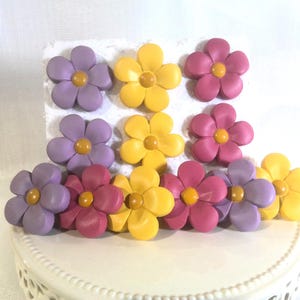 May include: A collection of eleven five-petaled flower decorations in shades of purple, yellow, and pink. Each flower features a small, round, mustard-yellow center. The flowers are arranged in a grid-like pattern on a white rectangular base.