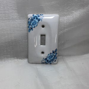 May include: A rectangular white ceramic light switch wall plate featuring a glossy finish and a blue floral pattern printed on the top left and bottom right corners. The plate includes a central vertical toggle switch cutout and two screw holes.
