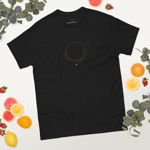 Puede incluir: Camiseta negra con un diseño circular dorado, una estrella y detalles punteados. La camiseta de cuello redondo está rodeada de frutas y vegetación. Un diseño sencillo y elegante.