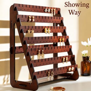 May include: A dark brown wooden earring display stand with multiple horizontal rows of slots for hanging earrings. The stand is angled and features the text "Showing Way". Several pairs of earrings are displayed on the stand.