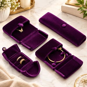 May include: A collection of deep purple velvet jewelry cases including a long rectangular box, a square box containing a gold butterfly necklace, a small ring box with two gold rings, and a square box holding a gold bangle bracelet.