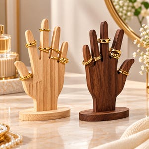 May include: Two hand-shaped jewelry displays made of wood, one light-toned and one dark-toned, each featuring an oval base. Both displays are adorned with multiple gold-colored rings worn on the fingers, showcasing a variety of band styles.