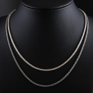 May include: Two silver-coloured chain necklaces displayed on a black mannequin. The necklaces have a woven design, with one chain appearing lighter in colour than the other. The chains are draped in a curved shape.