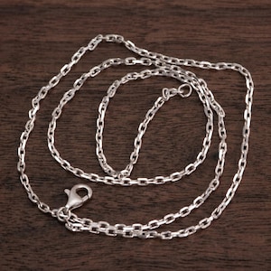 May include: A silver-colored chain necklace with an oval link design. The necklace has a lobster clasp closure. The chain is displayed on a dark brown wooden surface.