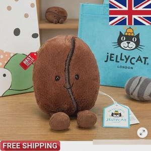 Könnte beinhalten: Ein braunes, plüschiges Kaffeebohnen-Spielzeug mit einem lächelnden Gesicht und kleinen Füßen. Ein "Best Seller"-Etikett hängt daran. Im Hintergrund ist eine blaue Tragetasche mit einem Katzenlogo und dem Text "JELLYCAT LONDON" zu sehen.