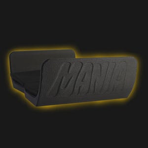 May include: A black, U-shaped holder with a textured surface and the word MANIA embossed in large, capital letters on the front panel. The object features a wide, open design with a flat base and two upright sides, surrounded by a soft yellow glow.