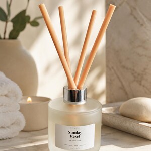 May include: A frosted glass diffuser bottle with a silver collar and light brown reeds. The label reads "Sunday Reset". The bottle is surrounded by a candle, a vase with greenery, and a rolled towel. The overall theme is relaxation and home fragrance.